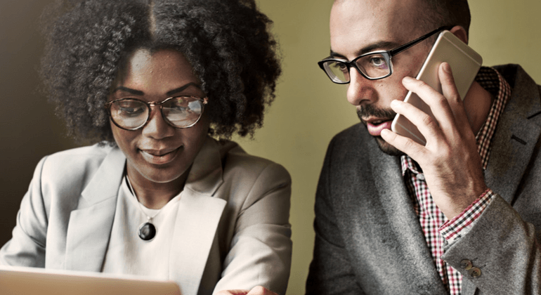 Man on cell phone sits with woman on a laptop as they collaborate 